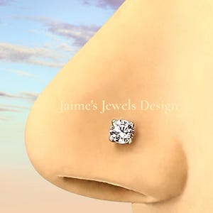 May include: A close-up of a nose with a square-cut, clear gemstone nose stud. The stud is set in a silver or white metal. The text "Jaime's Jewels Design" is visible in the background.