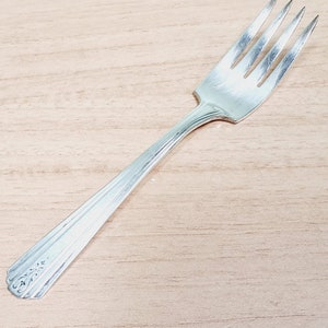 May include: A silver fork with four tines, resting on a light wood surface. The handle has a decorative floral design at the end and a series of parallel lines. The fork is a classic piece of silverware.
