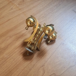 May include: Ornate gold-colored metal object with a complex, abstract design. Features multiple spherical elements and curved, stylized shapes. The object appears to be a decorative piece or part of a larger item, possibly vintage.