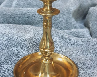 May include: A vintage brass candlestick holder with a round base and a fluted stem. The candlestick is gold-colored and has a classic design. It is likely used for holding a single candle. The candlestick is on a gray surface.