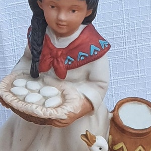 May include: A ceramic figurine of a young girl in a white dress, holding a basket of eggs. She has a red scarf and a braid. A white duck and a brown jug are nearby. The figurine is on a green base.