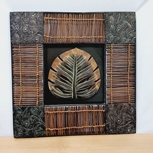 May include: Square wall art featuring a central leaf design in shades of brown and green. The leaf is set against a black background and framed by woven bamboo and decorative metal panels with floral patterns. A rustic, nature-inspired decor piece.