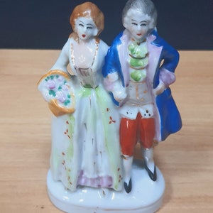 May include: A porcelain figurine depicting a couple in 18th-century attire. The woman wears a white dress with a green overskirt and carries a basket of flowers. The man is in a blue coat and red breeches. The figurine is on a white base.