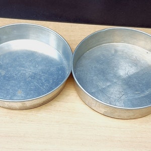 May include: Two round, silver-colored baking pans. The pans are made of metal and have a slightly scratched surface. The pans are empty and ready for use. The pans are sitting on a light brown wooden surface.