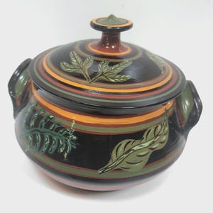 May include: A round ceramic lidded pot featuring a glossy black base with horizontal bands of orange and green. The exterior and lid are decorated with painted green leaf motifs. The pot includes two side handles and a central knob on the lid.