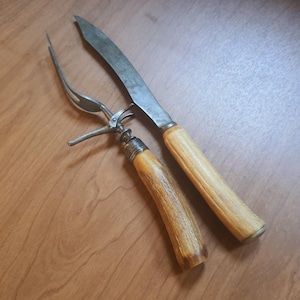 May include: Antique carving set featuring a knife and fork. The knife has a long, slightly curved blade and a light-colored handle. The fork has two tines and a matching handle. Both handles have a textured appearance and are made of a light brown material.