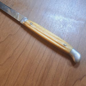 May include: A close-up of a knife with a light-colored wooden handle and a silver blade. The handle is secured with small rivets and has a curved, silver end. The knife is on a wooden surface.