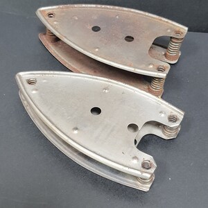 May include: Two antique flat irons, featuring a curved, silver-colored metal design with spring mechanisms. The irons show signs of age with rust and wear, suggesting they are vintage or collectible items.