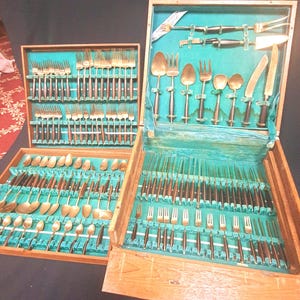 May include: A large flatware set housed in two wooden storage cases with teal velvet lining. The set includes gold-toned metal forks, spoons, and knives with dark brown handles, arranged in organized slots within the wooden boxes.