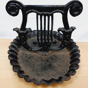 May include: Black cast iron decorative dish with a lyre design. The dish has a scalloped edge and a detailed lyre shape at the top. The dish is on a light wood surface.