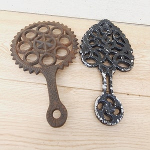 May include: Two cast iron trivets. One is rusty brown with a circular design and handle. The other is black with white accents and a tree-like design. Both trivets have a handle for easy use and are placed on a wooden surface.