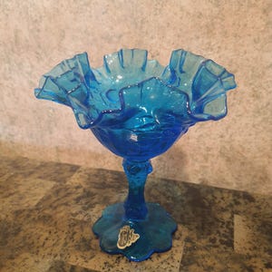May include: A vibrant blue glass pedestal bowl with a ruffled rim. The bowl sits on a stemmed base with a decorative, scalloped foot. The glass has a textured appearance and a small sticker is affixed to the base.