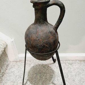 May include: A terracotta vase with a brown glaze and a handle, sitting on a black metal stand with three legs.