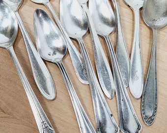 May include: A collection of antique silver spoons with various handle designs, including floral and geometric patterns. The spoons are arranged on a light wooden surface, showcasing their reflective, polished surfaces. Some spoons have engraved initials.
