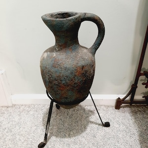 May include: A blue and brown ceramic vase with a handle, sitting on a black metal stand with three legs.