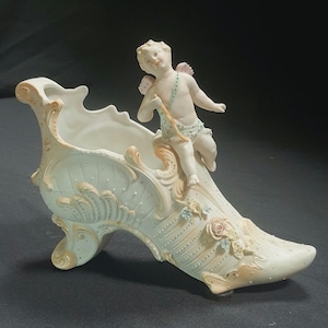 May include: A decorative porcelain shoe-shaped vase in off-white, adorned with floral and scrollwork details. A cherub figure with pink wings and a bow sits atop the shoe. The vase has a high heel and a detailed design.