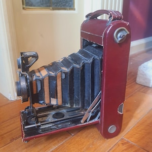 May include: Vintage burgundy leather camera with a black bellows and a handle. The camera has a metal lens and various mechanical components. The camera is set on a wooden floor.