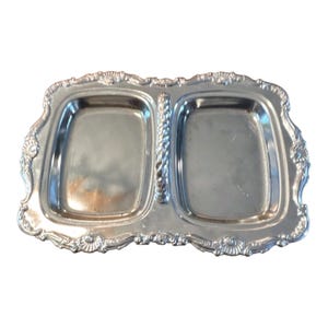 May include: A silver-toned metal serving tray featuring two rectangular compartments separated by a central vertical handle with a twisted rope texture. The tray has a decorative scalloped border with embossed shell-like patterns along the edges.