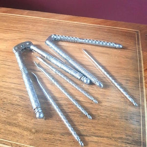 May include: A set of silver-toned metal nutcrackers and seafood picks featuring intricate, textured patterns along the handles. The collection includes one hinged nutcracker tool and four slender, pointed implements with decorative, grooved detailing.