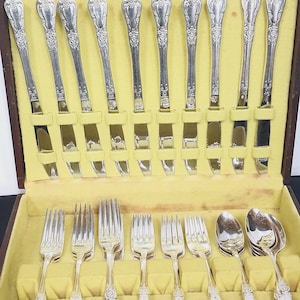May include: A silver-colored flatware set in a wooden case with a yellow interior. The set includes knives, forks, and spoons, all with ornate handle designs. The case is open, displaying the arranged silverware.