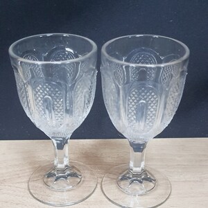 May include: Two clear, stemmed wine glasses with a detailed, textured pattern. The glasses have a rounded bowl shape and sit on a circular base. The background is a dark color, and the glasses are on a light-colored surface.