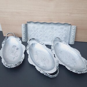 May include: A set of four silver-toned metal serving pieces including one rectangular tray and three oval-shaped bowls. Each piece features a hammered texture and embossed floral patterns, with decorative handles on the oval bowls.