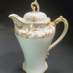 May include: A white porcelain coffee pot with a lid and handle. The pot is decorated with gold floral patterns and trim. The handle and lid finial are also gold. The pot has a curved, elegant shape.
