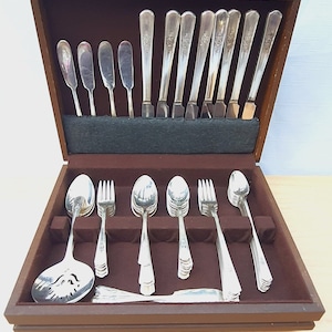 May include: A complete set of silver-colored flatware, including spoons, forks, and knives, neatly arranged in a wooden storage box. The flatware features a floral design on the handles. The box is lined with a dark brown fabric.