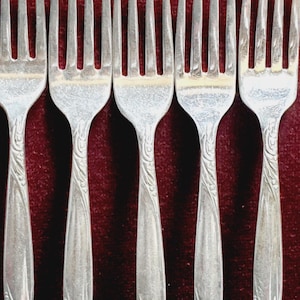 May include: Six silver forks with ornate floral designs and a monogram on the handle. The forks are arranged in a row on a red velvet background.
