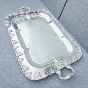May include: A rectangular silver serving tray with ornate handles and a decorative border featuring grape clusters. The tray has a slightly textured surface and is designed for serving food or beverages. The handles are curved and detailed.