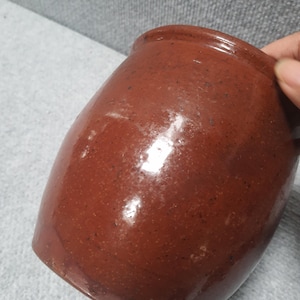 May include: A reddish-brown ceramic jar with a glossy finish. The jar has a rounded body and a slightly flared rim. The surface has subtle variations in color and texture, suggesting a handmade or antique quality. The jar is held by a hand.