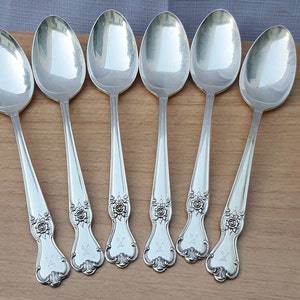 May include: Six silver spoons arranged on a wooden surface. Each spoon has an oval bowl and a long handle with a floral design and the letter "K". The spoons reflect the light, creating a bright, shiny appearance. The spoons are likely part of a silverware set.