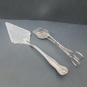 May include: A silver-toned metal cake server and a pair of matching sugar tongs. The triangular cake server features an intricate floral engraving on the blade and a shell motif on the handle. The tongs have scissor-style handles and ornate details.