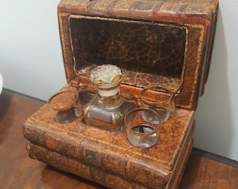 May include: A vintage-style brown leather book-shaped box with gold-toned accents, containing a clear glass decanter with a faceted stopper and three matching small glass tumblers, each featuring a thin gold rim around the top edge.