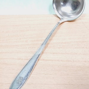 May include: A silver ladle with a round bowl and a long, decorated handle. The handle has a beaded edge and an engraved monogram. The ladle is made of a shiny, reflective metal and is set against a light wood surface.