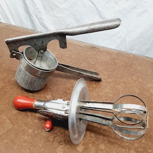 May include: Two vintage kitchen tools: a potato ricer and a hand-crank egg beater. The potato ricer is made of metal with a perforated bowl and a long handle. The egg beater has a red handle and a circular metal plate.