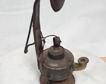 May include: An antique, rustic oil lamp with a circular reflector and a handle. The lamp is made of aged, dark metal with a small spout and a decorative fork-like tool hanging from a chain. The lamp sits on a white surface.