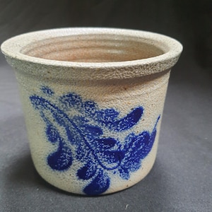 May include: A small, cylindrical stoneware pot with a light beige exterior and a dark blue floral design. The rim and interior are a lighter beige color. The pot is likely used for planting or storage.
