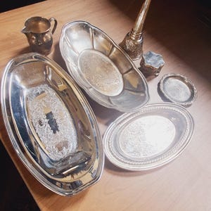 May include: An assortment of antique silver serving pieces, including oval platters, a small creamer, a small dish, and a decorative candle holder. The pieces are arranged on a light brown wooden surface, reflecting the surrounding light.