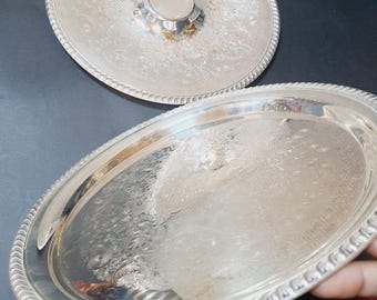 May include: Two silver-colored serving trays. One is oval-shaped, and the other is round with a small, raised center. Both trays have a decorative, rope-like border and etched floral patterns. The trays are reflective and appear to be made of metal.