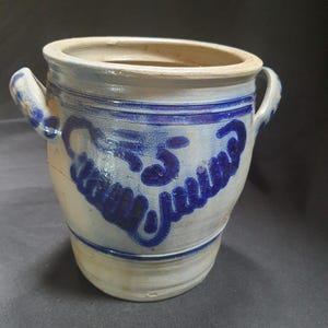 May include: A vintage stoneware crock with a blue and white design. The crock has two handles and a blue painted inscription. The exterior is a light beige color with blue accents. The crock is a cylindrical shape.