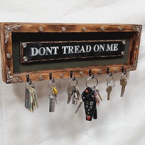 May include: Wooden key holder with the text "DONT TREAD ON ME". The key holder has a dark brown frame with decorative metal accents and hooks for keys. Various keys and keychains hang from the hooks.