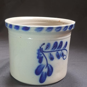 May include: A white ceramic crock with a blue floral design. The rim features a series of blue dots. The side of the crock has a blue leaf and vine pattern. This vintage-style container is suitable for kitchen or home decor.