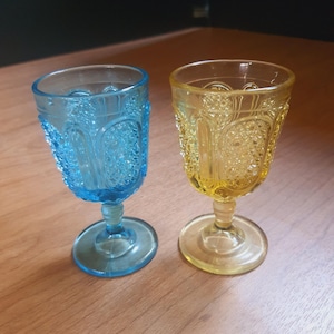 May include: Two vintage-style, stemmed glasses. One is a vibrant blue, the other a sunny yellow. Both glasses have a detailed, embossed pattern around the bowl and a circular base. They sit on a wooden surface.