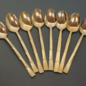 May include: A set of seven gold-colored spoons arranged on a dark surface. The spoons have a bamboo-style handle design and a polished, reflective surface. The spoons are likely for serving or eating.