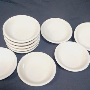 May include: A collection of small, off-white ceramic dishes. Some are stacked, while others are arranged around the stack. The dishes have a simple, round design with slightly raised edges. They are set against a dark background.