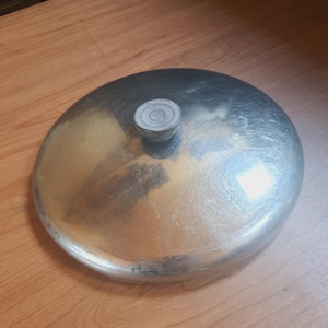 May include: A round, silver-colored metal lid with a small, circular handle on top. The lid has a slightly domed shape and shows signs of wear. The surface reflects light, indicating a polished finish. This item is likely a kitchenware accessory.