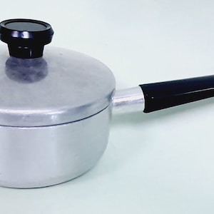 May include: A small, silver-colored saucepan with a black handle and lid. The lid has a black knob. The handle is approximately 6 inches long. The saucepan is designed for cooking and is likely made of aluminum.