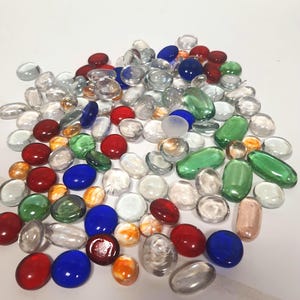 May include: A collection of smooth, rounded glass decorative stones in various colors including clear, red, blue, green, and orange. These flat-bottomed glass gems vary in size and shape, featuring a polished, reflective surface throughout the pile.