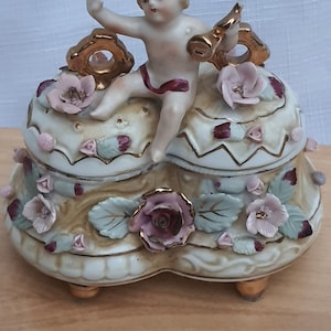 May include: A porcelain trinket box with a cherub figurine on top. The box is heart-shaped, cream-colored, and decorated with pink roses, gold accents, and green leaves. The cherub is nude with blonde hair and a red sash.